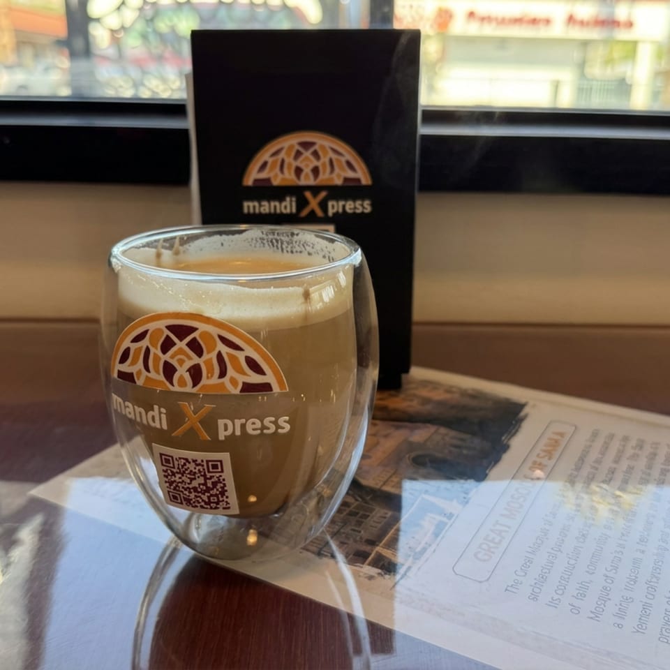 Mandi Xpress-branded frothy drink sits on a glossy restaurant table beside a menu and window.