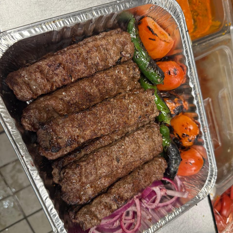 Grilled minced-meat kebabs arranged in a foil tray with charred tomatoes, green peppers, black eggplant, and sliced red onions.