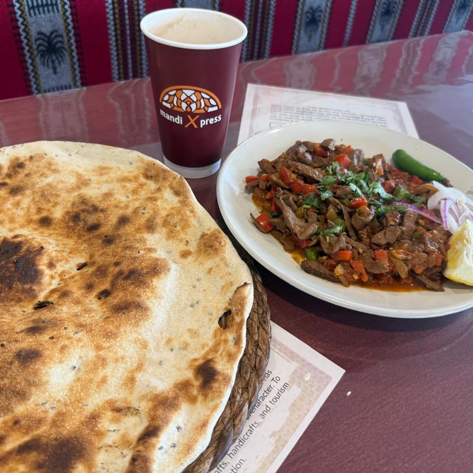 Mandi Xpress meal with grilled flatbread, spiced meat and vegetables, lemon, onions, chili, and a cup on a restaurant table.