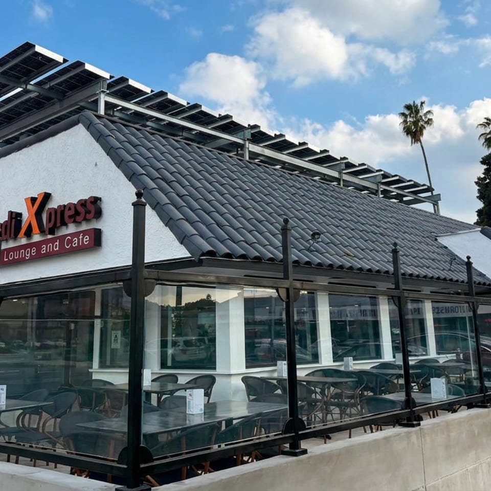 Exterior of macchi Xpress Yemeni lounge and cafe with glass-enclosed patio, tiled roof, solar panels, and palm trees.