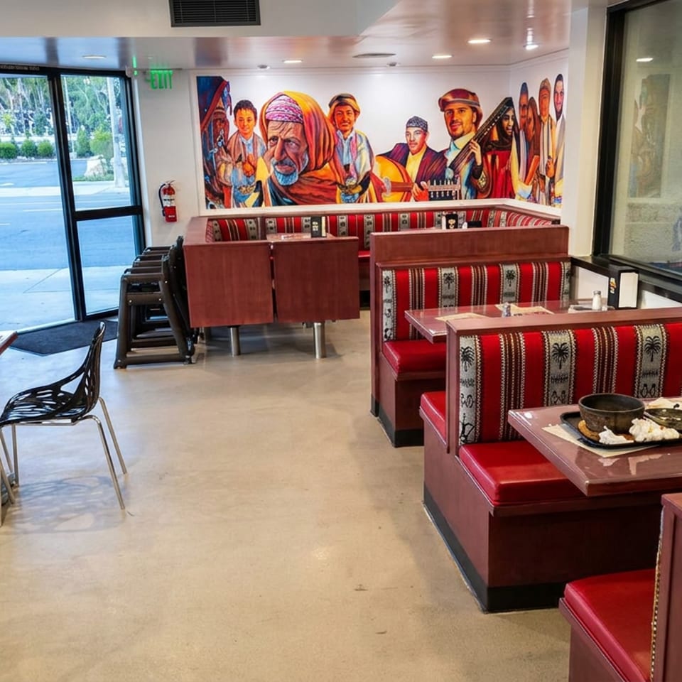 Bright restaurant dining room with red patterned booths, black chairs, tables, and a colorful mural of Middle Eastern figures.
