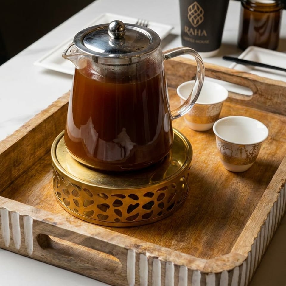 Glass pot of dark tea on a brass warmer, accompanied by small patterned cups on a wooden serving tray.