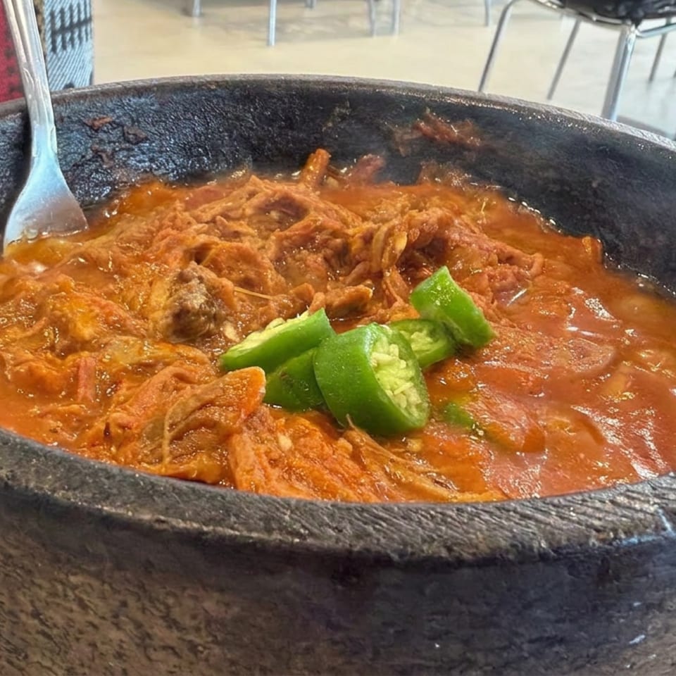 Shredded meat simmered in red sauce with lime wedges, served in a black stone bowl on a restaurant table.