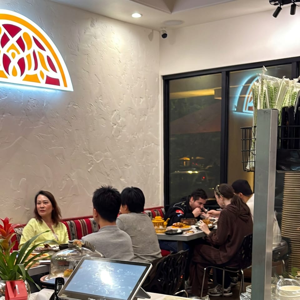 Restaurant interior with diners enjoying meals beneath an illuminated red-and-gold logo, while stacked plates and utensils occupy the foreground.