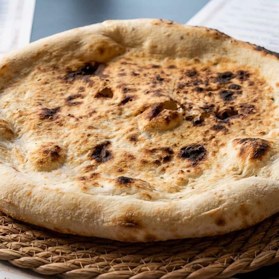 Round, blistered flatbread with a thick golden crust rests on a woven basket plate atop a restaurant table.